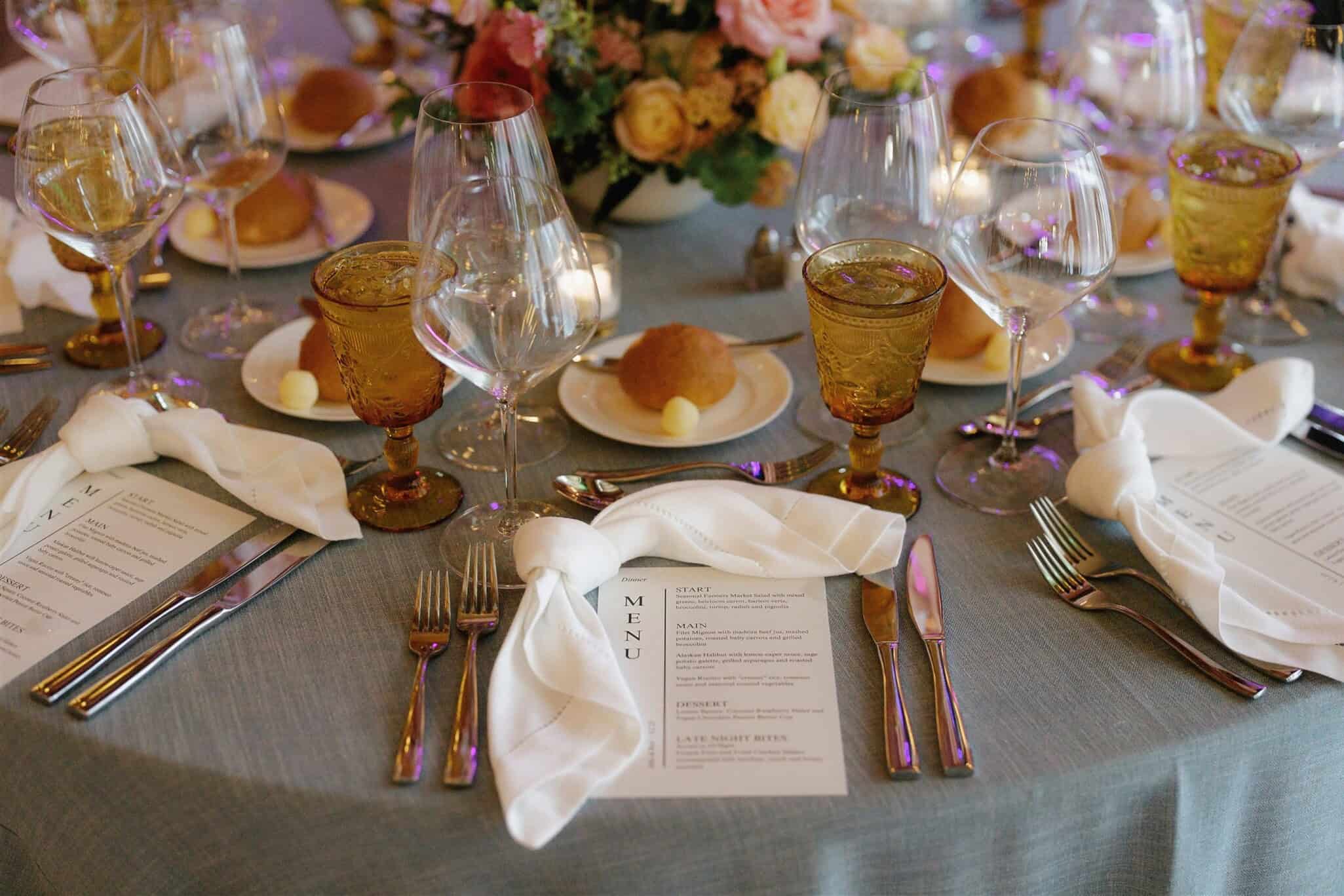 Formal dinner table setting with menus, gold-toned glasses, folded white napkins, bread rolls, butter, cutlery, and a floral centerpiece on a gray tablecloth.
