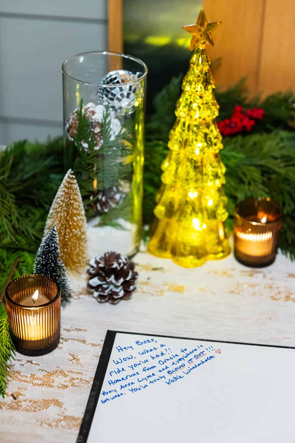 A tabletop decorated with lit candles, evergreen branches, pinecones, artificial trees, and a glowing yellow Christmas tree. A handwritten note is visible.