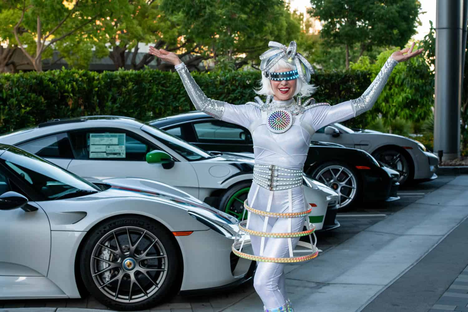 Woman in a futuristic white costume and wig poses with arms raised in front of a row of parked sports cars outdoors.