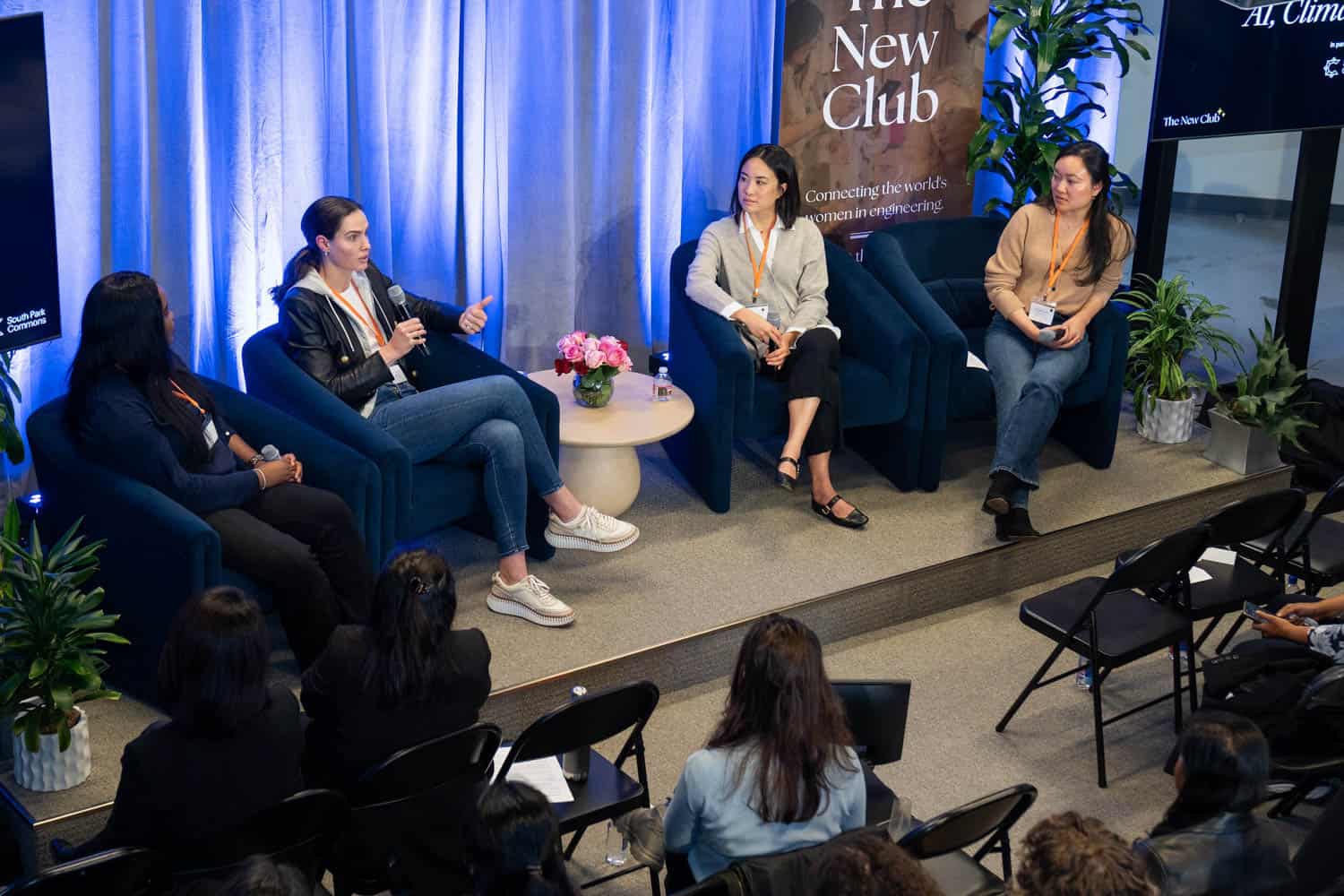 Four women sit on a stage panel discussing, with an audience watching. Banners and a screen display The New Club and technology themes.